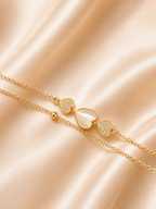 A delicate gold chain bracelet with three heart charms, featuring two pavé-set heart charms and one larger heart with a milky white inlay, displayed on a draped champagne satin fabric. The Whispering Hearts Bracelet by Ashaarya featuring 18KT gold plating, mother of pearl hearts, cubic zirconia, anti-tarnish 316L stainless steel comfort jewellery.