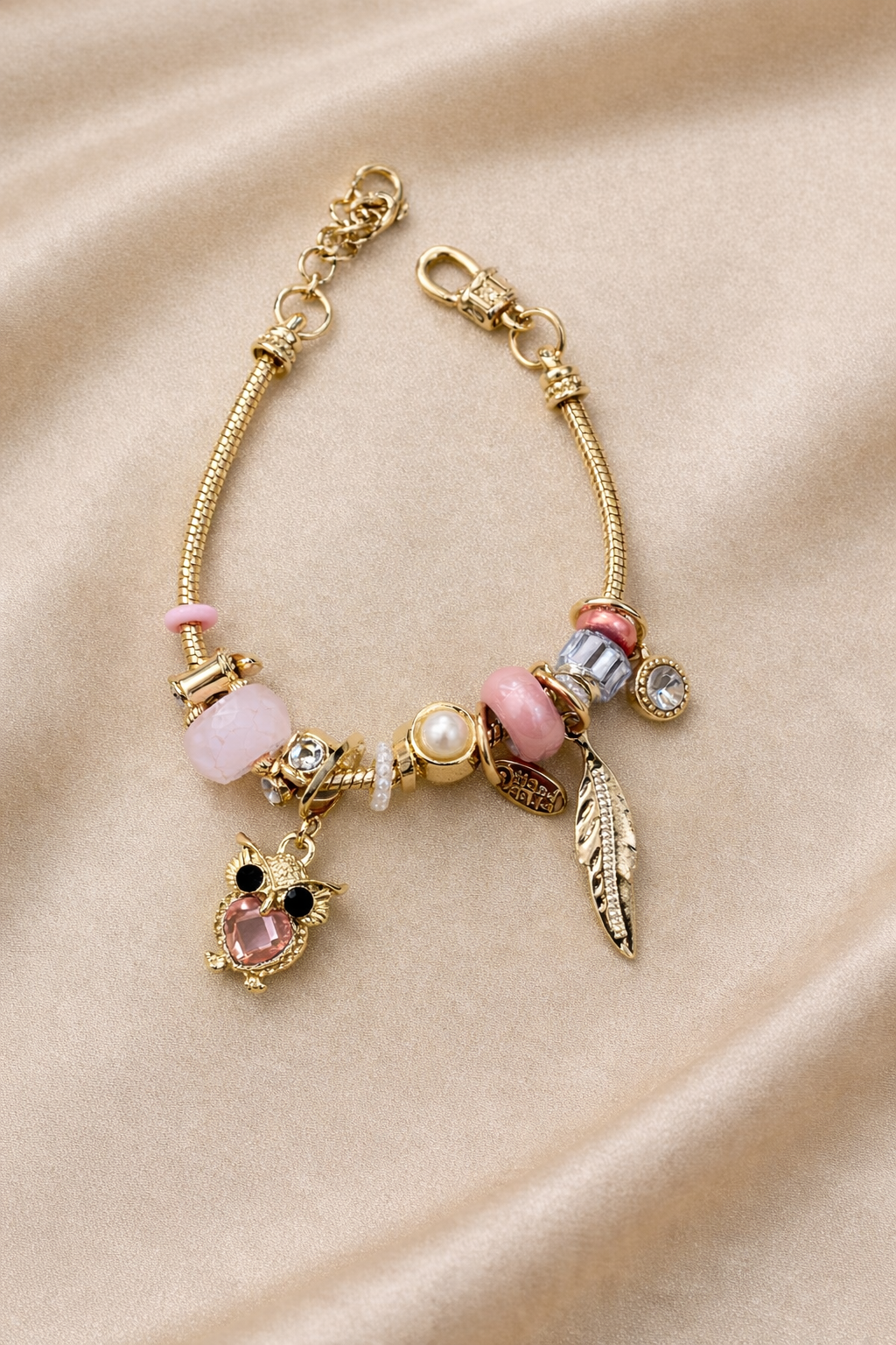 Anti-tarnish pink owl charm bracelet with feather and crystal charms on gold snake chain – comfort jewellery by Ashaarya. Gold bracelet with various charms on a beige fabric background