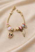 Anti-tarnish pink owl charm bracelet with feather and crystal charms on gold snake chain – comfort jewellery by Ashaarya. Gold bracelet with various charms on a beige fabric background