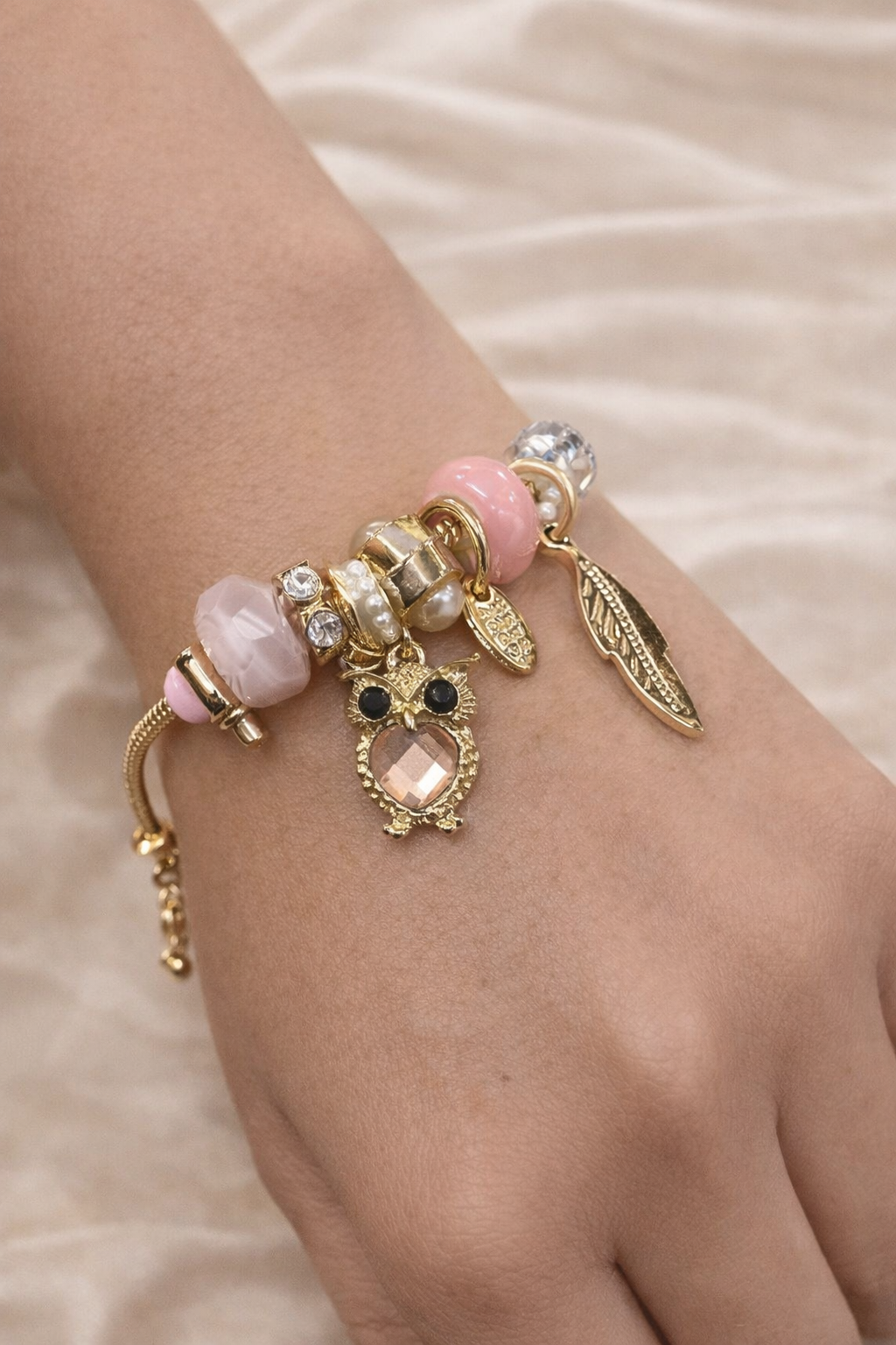 Anti-tarnish pink owl charm bracelet with feather and crystal charms on gold snake chain – comfort jewellery by Ashaarya. Delicate bracelet with pink beads and gold charms on a wrist against a soft background