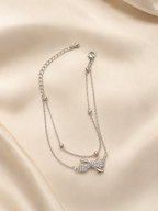 A delicate silver double-strand chain anklet featuring a small bow charm encrusted with clear stones, displayed on a draped cream satin fabric background. Bow of Grace Bracelet by Ashaarya – anti-tarnish silver bow bracelet with double chain and bead details Anti-tarnish Grace Bow Comfort Bracelet by Ashaarya crafted from 316L stainless steel with sparkling American Diamonds.