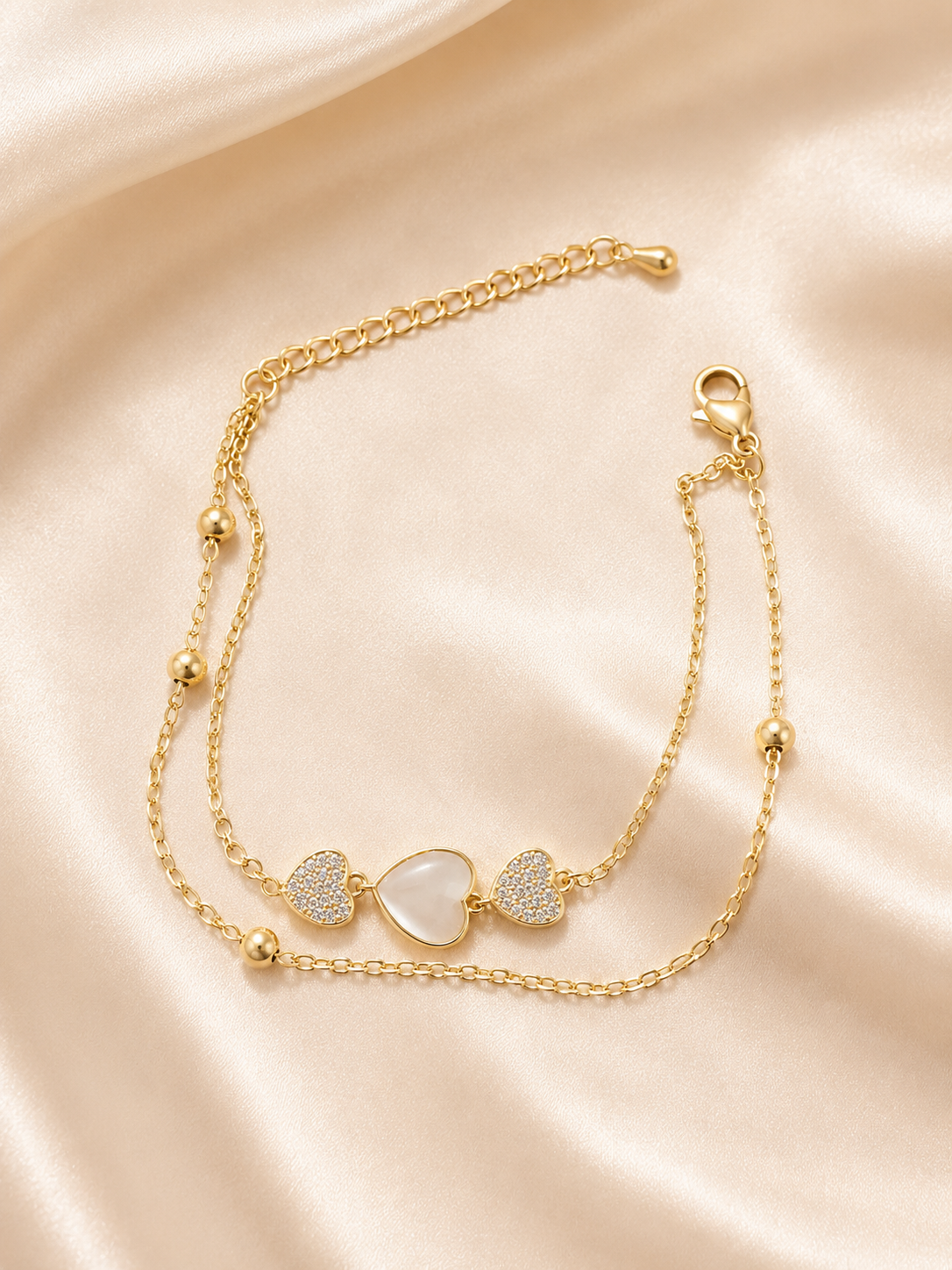 A gold double-layered anklet featuring three heart charms, including one central iridescent white heart and two textured side hearts, displayed on a draped beige satin fabric.