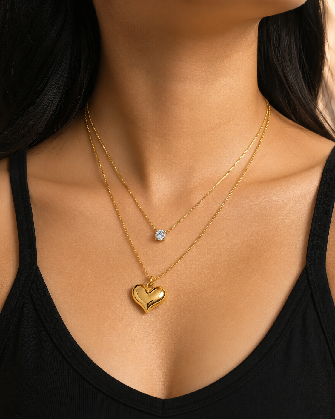 A close-up of a model wearing a double-layered gold necklace set. The top layer features a delicate chain with a small round clear gemstone pendant, while the bottom layer has a slightly thicker chain with a polished gold heart-shaped pendant. everyday heart necklace, heart necklace for women, layered necklace for women, gold heart necklace, minimalist gold necklace, anti tarnish necklace, waterproof necklace, 18kt gold plated necklace, stainless steel necklace, dainty layered necklace, everyday jewellery, 