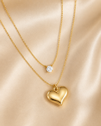 Two delicate gold necklaces displayed on a soft champagne satin background. The shorter necklace features a small round clear gemstone pendant, while the longer necklace has a polished gold heart-shaped pendant. everyday heart necklace, heart necklace for women, layered necklace for women, gold heart necklace, minimalist gold necklace, anti tarnish necklace, waterproof necklace, 18kt gold plated necklace, stainless steel necklace, dainty layered necklace, everyday jewellery, lightweight necklace, comfort je