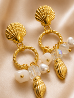 A pair of gold-toned statement earrings with scalloped shell-shaped studs, twisted rope rings, faux pearl beads, and small iridescent crystal drops, displayed on a soft beige satin fabric background. statement earrings for women, shell earrings, pearl drop earrings, beach inspired earrings, gold statement earrings, anti tarnish earrings, waterproof earrings, 18kt gold plated earrings, stainless steel earrings, coastal jewellery, lightweight earrings, comfort jewellery, vacation earrings, luxury earrings for