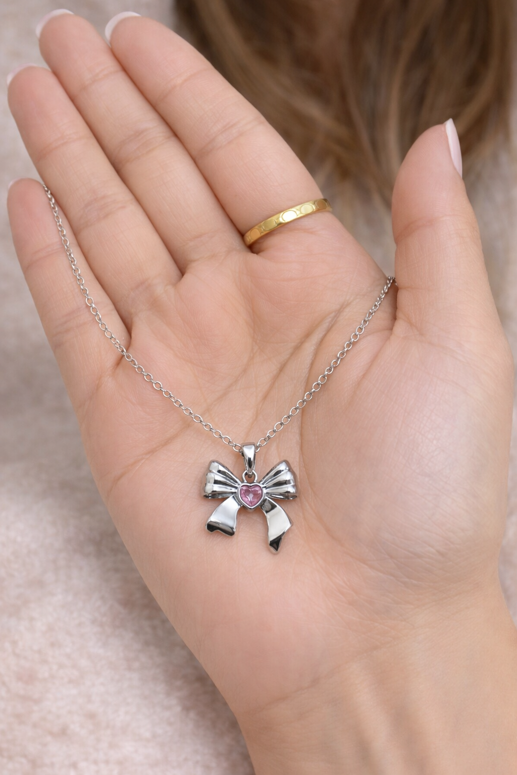 Anti tarnish necklace, Ashaarya, Charm bracelets, Ashaarya Jewels, Ashaarya jewellery, Hand holding a silver necklace with a bow pendant featuring a pink heart.