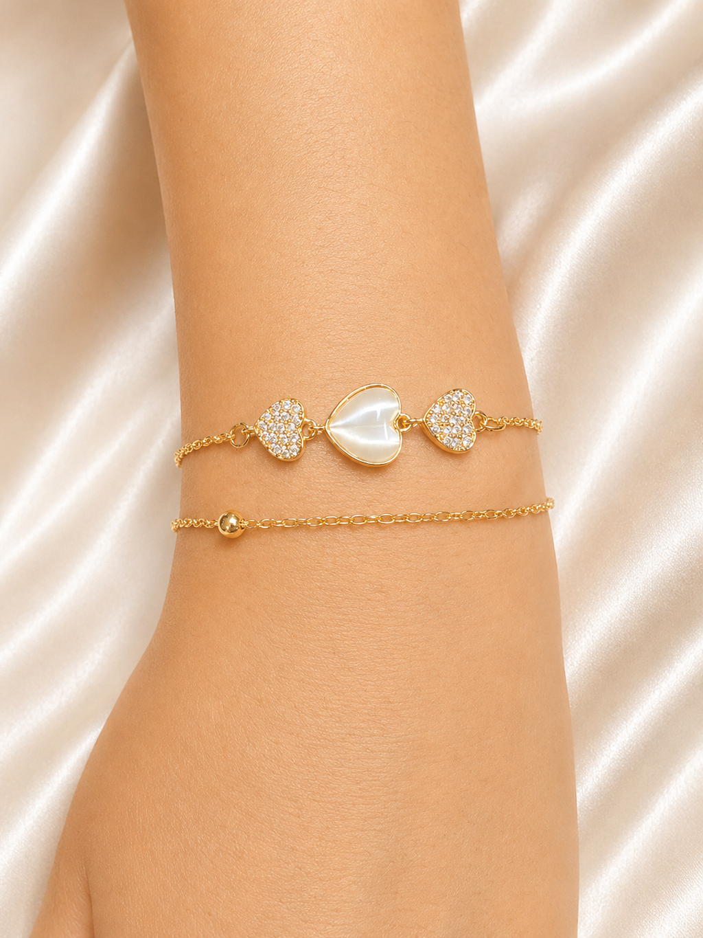 A close-up of a person's wrist wearing two delicate gold bracelets. The top bracelet features a central white mother-of-pearl heart flanked by two pavé-set heart charms. The Whispering Hearts Bracelet by Ashaarya featuring 18KT gold plating, mother of pearl hearts, cubic zirconia, anti-tarnish 316L stainless steel comfort jewellery.
