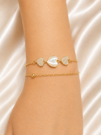 A close-up of a person's wrist wearing two delicate gold bracelets. The top bracelet features a central white mother-of-pearl heart flanked by two pavé-set heart charms. The Whispering Hearts Bracelet by Ashaarya featuring 18KT gold plating, mother of pearl hearts, cubic zirconia, anti-tarnish 316L stainless steel comfort jewellery.