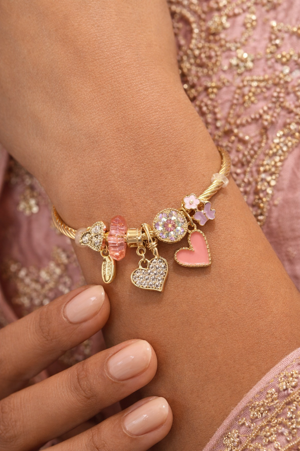 Gold bracelet with heart-shaped charms on a wrist against a pink embroidered background