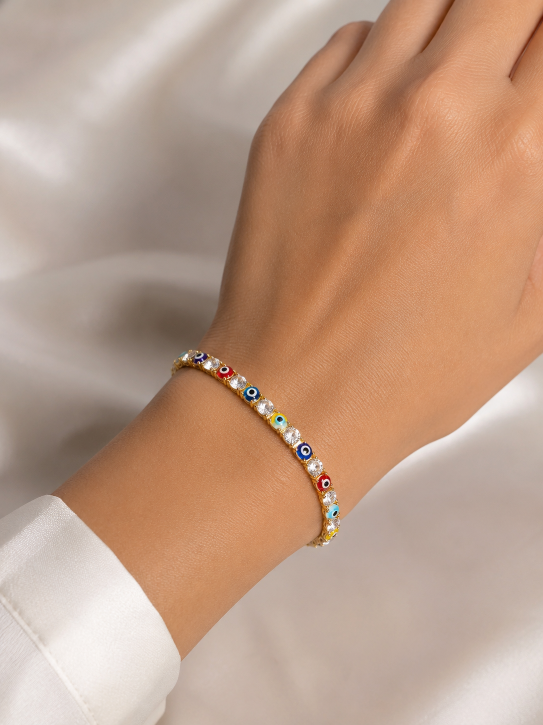 A delicate beaded bracelet with colorful evil eye charms and white pearl-like beads, shown worn on a person's wrist against a soft white satin background.