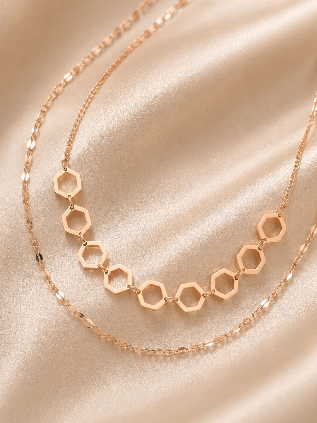 A rose gold double-layer necklace featuring a top layer with seven connected hexagonal openwork links and a bottom layer of a delicate cable chain, displayed on a draped champagne satin fabric. rose gold necklace for women, dual layer necklace, layered necklace for women, geometric necklace, honeycomb necklace, minimalist necklace, rose gold jewellery, anti tarnish necklace, lightweight necklace, stainless steel necklace, everyday necklace, office wear necklace, dainty layered necklace, comfort jewellery, p