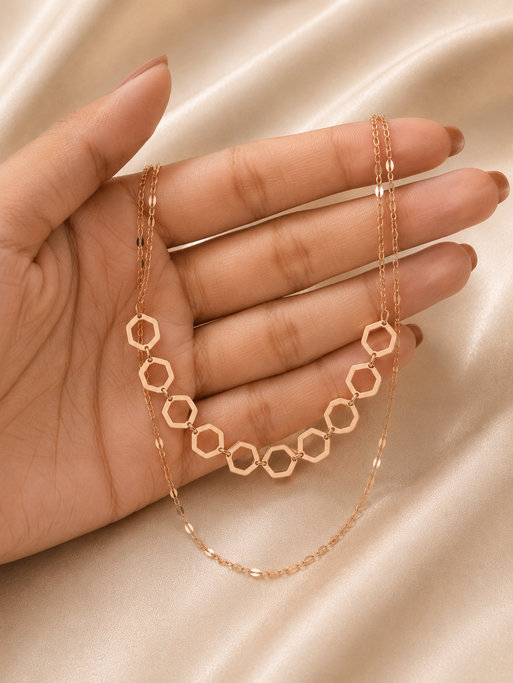 A rose gold double-layered necklace with one layer of hexagonal links and a thinner chain layer, displayed on a hand over a beige satin fabric background. rose gold necklace for women, dual layer necklace, layered necklace for women, geometric necklace, honeycomb necklace, minimalist necklace, rose gold jewellery, anti tarnish necklace, lightweight necklace, stainless steel necklace, everyday necklace, office wear necklace, dainty layered necklace, comfort jewellery, premium necklace for women, Ashaarya nec