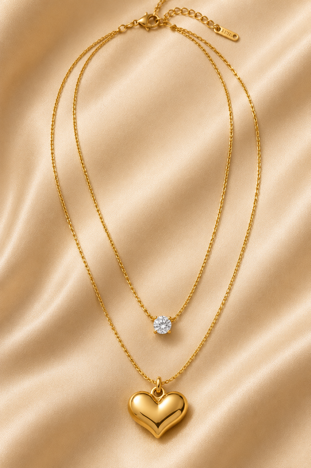 A double-layered gold necklace featuring a small round clear stone pendant on the upper layer and a larger polished gold heart pendant on the lower layer, displayed on a draped beige satin fabric background. everyday heart necklace, heart necklace for women, layered necklace for women, gold heart necklace, minimalist gold necklace, anti tarnish necklace, waterproof necklace, 18kt gold plated necklace, stainless steel necklace, dainty layered necklace, everyday jewellery, lightweight necklace, comfort jewell
