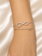 A delicate silver bracelet with an infinity symbol paved with clear stones, layered on a thin silver chain bracelet with small clear bead accents, shown on a model's wrist against a satin fabric background.
