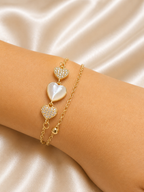 A gold chain bracelet with three heart charms worn on a person's wrist, set against a draped satin fabric background.