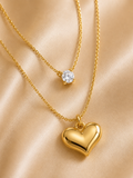 Two gold necklaces displayed on a satin fabric background. The shorter necklace features a single round clear gemstone pendant, while the longer necklace has a polished yellow gold heart-shaped pendant. everyday heart necklace, heart necklace for women, layered necklace for women, gold heart necklace, minimalist gold necklace, anti tarnish necklace, waterproof necklace, 18kt gold plated necklace, stainless steel necklace, dainty layered necklace, everyday jewellery, lightweight necklace, comfort jewellery, 