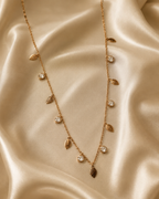 A gold leaf necklace with clear crystal accents, draped elegantly on a shimmering beige satin fabric background. rose gold necklace for women, anti tarnish necklace, leaf pendant necklace, nature inspired necklace, lightweight necklace for women, stainless steel necklace, 18kt rose gold necklace, dainty necklace for women, everyday necklace, minimalist jewellery, comfort jewellery, waterproof necklace, skin friendly necklace, Ashaarya necklace
